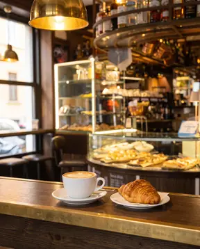 Un cappuccino e un cornetto sul banco del bar, con la vetrina dei salati dietro